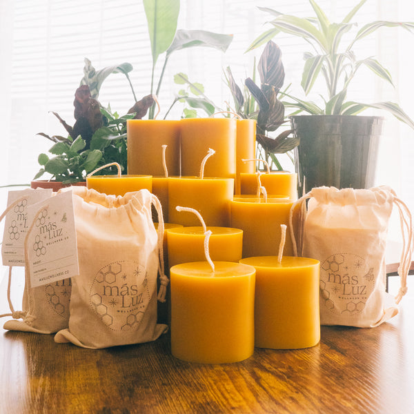 a group of various of beeswax pillar candles in various sizes. some of the beeswax pillars are packaged in a stamped drawstring cotton bag. in the background are various potted house plants