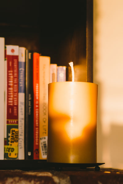 3" by 5" beeswax pillar candle in front of books on a shelf with shadows casting on it.